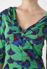 Grön blommig klänning med en vriden halsringning, rynkad textur och ett livfullt mönster av blå och röda blommor mot en grön bakgrund.