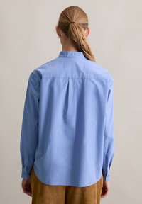 Vrouw met een blonde paardenstaart, gekleed in een losse lichtblauwe langemouwen shirt en een bruine broek, met haar rug naar de camera gericht tegen een effen beige achtergrond.