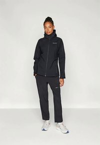 Femme se tenant debout, portant une veste zippée noire et un pantalon noir avec des baskets blanches, devant un fond clair et uni.