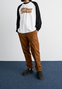Weißes und schwarzes Langarmshirt mit "BROWN DUCKS"-Grafik; braune Hosen mit Nike-Logo; schwarze Clogs mit flacher Sohle und erhöhter Ferse.