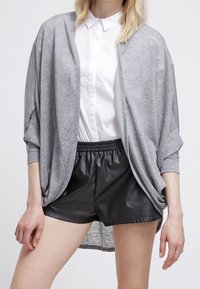 Cardigan oversized grigio chiaro con maniche a pipistrello, indossato sopra una camicia bianca con colletto e pantaloni corti in pelle nera. Tessuto morbido, design casual.