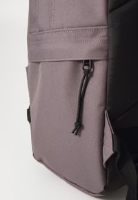 Purple fabric backpack with a textured surface, featuring a black accent, a drawstring closure, and a side pocket for additional storage.