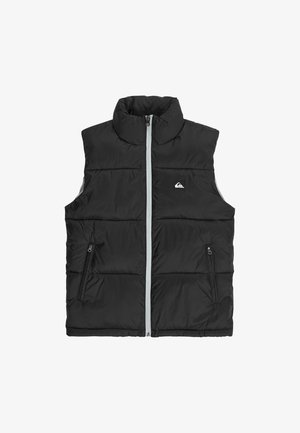 Niño sonriente que lleva un gorro rojo, chaleco acolchado negro sobre una sudadera negra, jeans azules holgados y zapatillas marrones, de pie contra un fondo blanco.