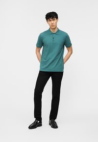 Jeune homme portant un polo à manches courtes couleur sarcelle, un pantalon noir et des chaussures habillées noires, debout les mains derrière le dos sur un fond uni.