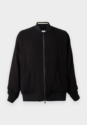 Blouson aviateur noir à fermeture éclair, avec col, poignets et ourlet côtelés, doté de deux poches avant, sur un fond gris clair uni.