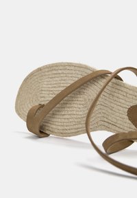 Bruine suède sandaal met een gevlochten jute zool, voorzien van twee slanke banden en een platte, gestructureerde zool voor grip en comfort.