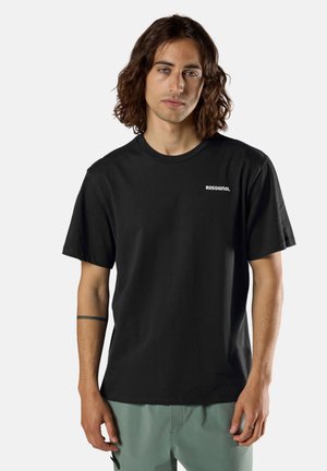 Młody mężczyzna z brązowymi włosami do ramion, ubrany w czarną koszulkę Rossignol i zielone spodnie na jednolitym tle.