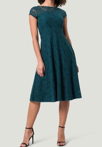 Robe en dentelle teal à manches courtes, corps ajusté et jupe évasée, présentant un motif de feuilles et une texture lisse, associée à des talons noirs.