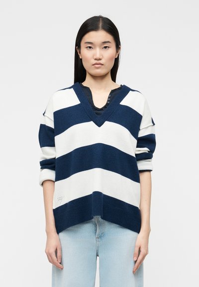 Jeune femme aux cheveux noirs raides portant un pull à col en V à larges rayures bleu marine et blanc, et un jean bleu clair, debout devant un fond uni.