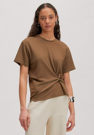 Frau mit langen lockigen Haaren trägt braunes T-Shirt mit verdrehtem Knoten, cremefarbene strukturierte Hose, silberne Creolen und ein breites silbernes Armreif.