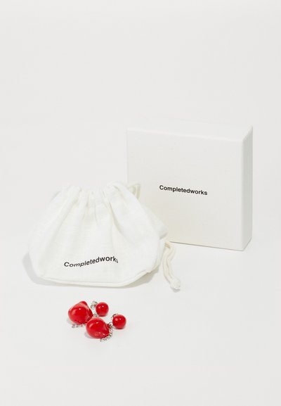 Borsa bianca con cordino riportante la scritta "Completedworks", contenente gioielli rossi. Accompagnata da una semplice scatola bianca.
