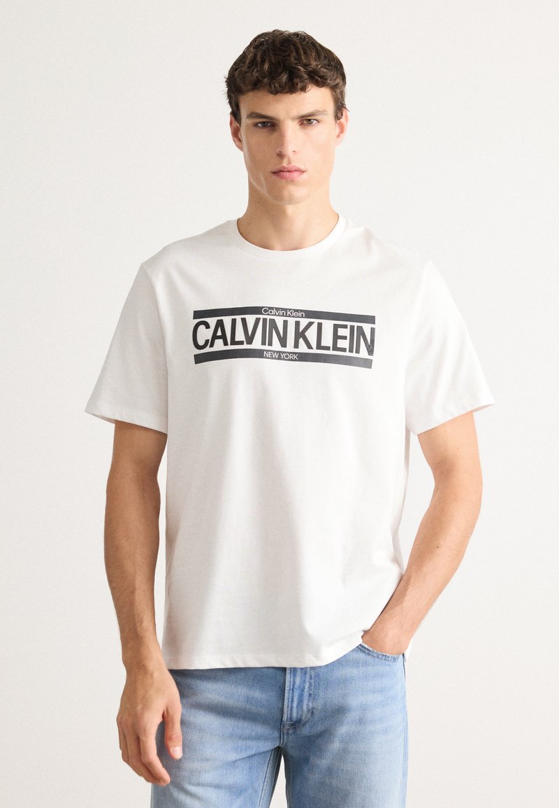 White cotton t-shirt featuring a black graphic print with "Calvin Klein" and "New York" text, short sleeves, and a crew neckline.