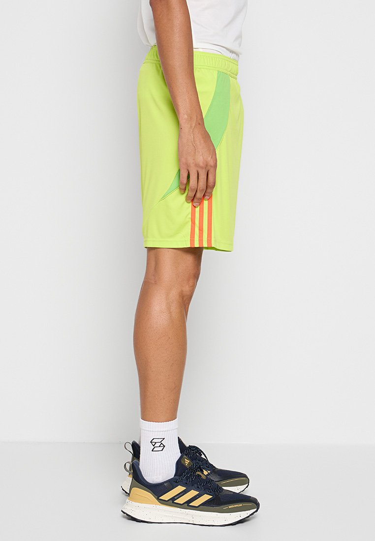 Shorts de sport vert lime avec poches latérales, dotés d'un panneau vert contrastant et de trois rayures orange sur le côté gauche. Tissu léger.