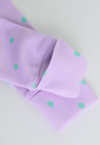 Tissu lilas avec des pois verts, montrant un détail de revers plié sur une manche de vêtement sur un fond uni.