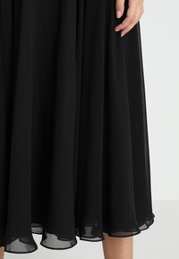 Jupe noire fluide avec des bords en tissu transparent, portée par une personne dont la main repose doucement sur le côté de la jupe.