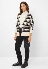 Stribet cardigan i creme og sort, med en bindebælte i taljen, åben front og teksturerede ærmer. Stylet med sorte bukser og kraftige sorte støvler.