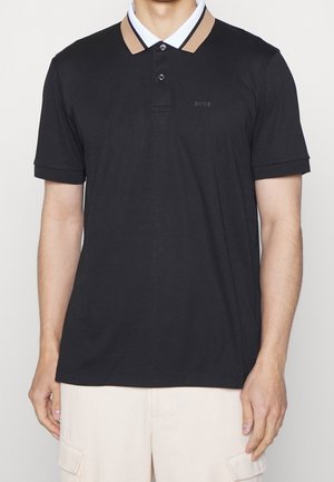 Homme portant un polo noir à manches courtes avec une bordure beige sur le col et le logo "BOSS" sur la poitrine, associé à un pantalon cargo de couleur claire.