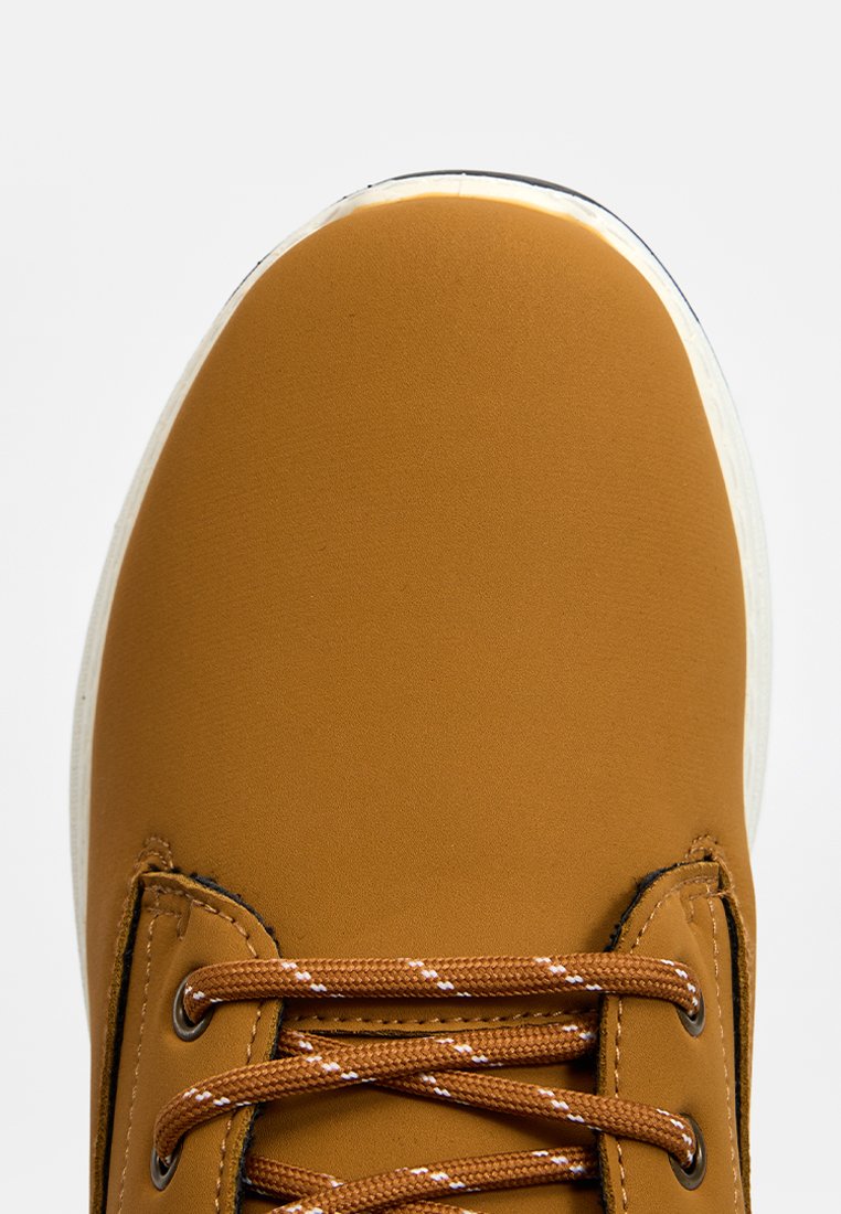 Braune Sneaker mit glattem Obermaterial, runder Zehenpartie, weißer Sohle und braunen Schnürsenkeln mit weißen Akzenten, die ein einfaches, modernes Design präsentieren.