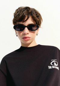T-shirt noir portant un graphique blanc du logo "Les Indestructibles". Le modèle porte des lunettes de soleil ovales noires et des boucles d'oreilles créoles, avec des cheveux courts et ondulés.