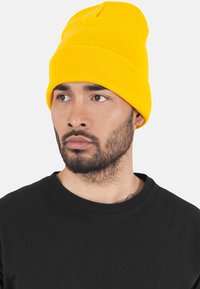 Yellow knit beanie with a cuffed design, made of soft fabric, featuring a seamless top and a vibrant color contrasting against a black shirt.