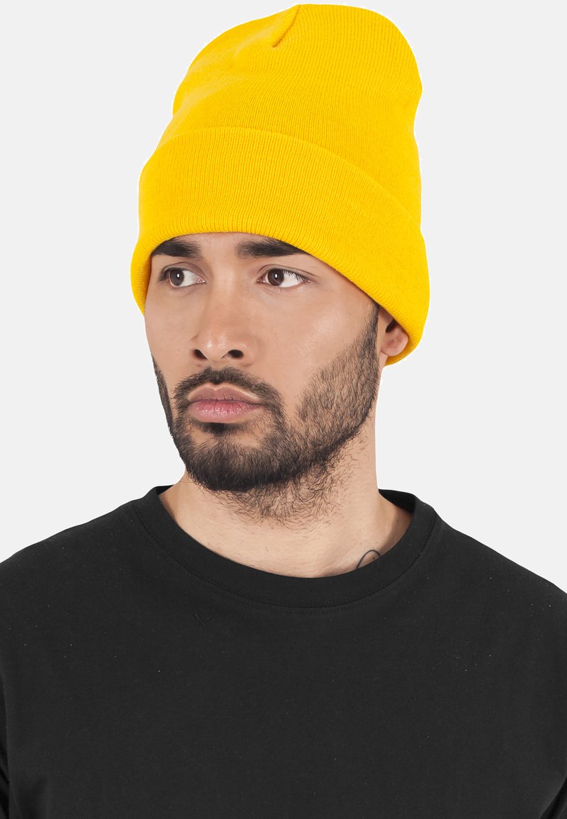 Yellow knit beanie with a cuffed design, made of soft fabric, featuring a seamless top and a vibrant color contrasting against a black shirt.