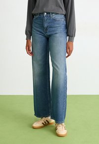 Person wearing high-waist wide-leg blue jeans, beige sneakers with brown stripes, and a long-sleeve dark gray top, standing on green floor.