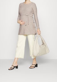 Strick-Tunika in Beige mit einem gegürteten Taillen Bereich und seitlichen Knöpfen, kombiniert mit hellen, knöchellangen Hosen und schwarzen Blockabsatz-Sandalen, die eine Beuteltasche in Beige hält.