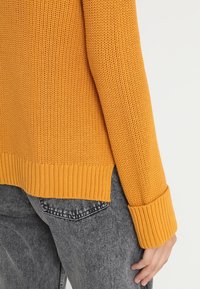 Gros plan sur une personne portant un pull en maille jaune moutarde avec des manches retroussées et un jean noir délavé, la main reposant à côté de son corps.