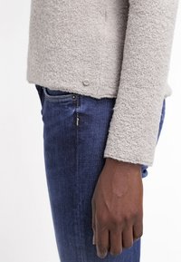 Main à la peau foncée reposant à côté d'un jean en denim bleu et d'un haut à manches longues gris clair texturé avec un petit détail bouton près de l'ourlet.