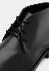 Μαύρο δερμάτινο παπούτσι με λεία, γυαλιστερή επιφάνεια, μαύρες κορδόνες και με κομμένο χώρο για τα κορδόνια. Ραμμένες λεπτομέρειες στην άκρη.