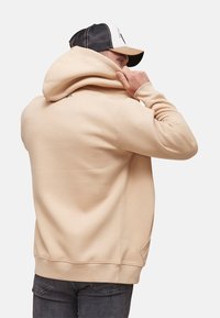 Sweat à capuche beige en matériau doux, avec une poche kangourou, des poignets côtelés et une capuche avec cordon de serrage. Vue arrière du porteur.