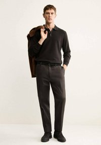 Polo shirt en tricot noir et pantalon gris foncé. Le modèle tient une veste marron. Les chaussures sont noires avec une finition texturée. Style simple, moderne.