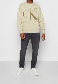 Sudadera beige con gran logo marrón "CK" y texto, combinada con jeans oscuros y zapatillas blancas. Cuenta con un cuello redondo y puños acanalados.