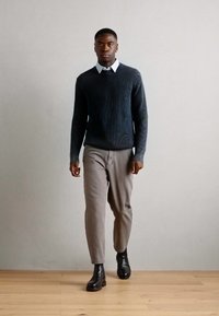 Un pull côtelé bleu foncé sur une chemise collée blanche, associé à un pantalon gris clair et des bottines noires, le tout sur un fond neutre.
