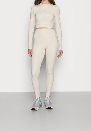 Woman wearing beige long-sleeve cropped top and high-waisted leggings with iridescent pastel sneakers, standing against a plain background.