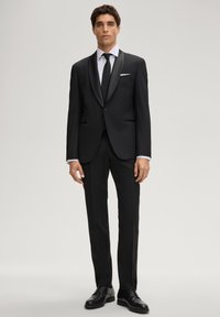 Schwarzer Smoking mit einem Satin-Schal-Kragen, Ein-Knopf-Verschluss und zwei Vorder Taschen. Kombiniert mit einem weißen Hemd und einer schwarzen Krawatte.