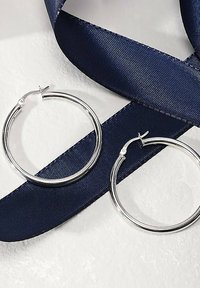 Orecchini a cerchio in argento con un design circolare liscio, appoggiati su un nastro di satin blu scuro su uno sfondo chiaro.