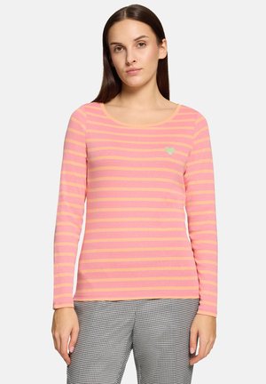 Woman wearing a pink and orange striped long-sleeve top with a small heart emblem on the chest, paired with black and white patterned pants.