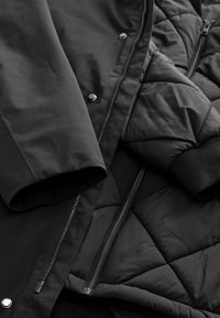 Gros plan d'une veste d'hiver noire montrant le tissu matelassé, la fermeture éclair, les boutons-pression et le poignet côtelé sur la manche.