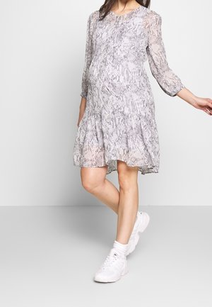 Kobieta w ciąży ubrana w długą, jasnoszarą sukienkę ciążową z wężowym wzorem i białe trampki, stojąca z jedną lekko uniesioną nogą.