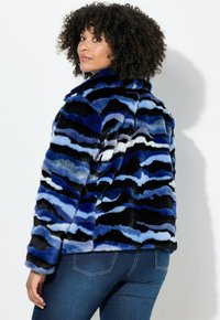 Veste en fausse fourrure avec un motif ondulé bleu et noir, un col montant et une coupe décontractée. Un jean en denim bleu est visible en dessous.