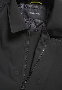 Manteau noir avec doublure matelassée, fermeture éclair partiellement ouverte, boutons-pression et une boucle jaune pour suspendre à l'intérieur du col.