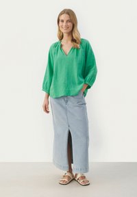 Grüne Leinenbluse mit V-Ausschnitt und gerafftem Kragen, kombiniert mit einem hellblauen Denim-Rock mit Schlitz vorne und beigen Sandalen.