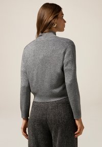 Maglione grigio corto con collo alto e orlo a coste, realizzato in un tessuto testurizzato con una leggera lucentezza, abbinato a pantaloni grigio scuro.