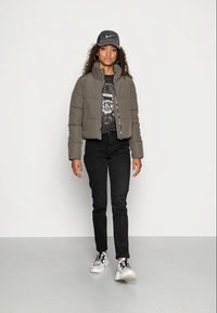Olive green puffer jacket, black graphic t-shirt, black jeans, and sneakers. Curly hair under a gray cap, standing against a plain background.
