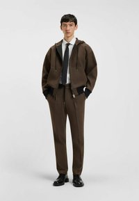 Bruine capuchonjack met ritssluiting aan de voorkant en zakken, gecombineerd met bijpassende bruine stijlvolle broek. Bevat zwarte accenten en een wit shirt.