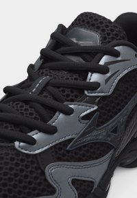 Μαύρο αθλητικό παπούτσι με ύφασμα από δίχτυ, μαύρα κορδόνια και ασημένιες συνθετικές επικαλύψεις με στυλιζαρισμένο λογότυπο βέλους στο πλάι.