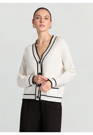 Donna che indossa un cardigan bianco con bordi e bottoni neri, abbinato a pantaloni neri, in piedi contro uno sfondo chiaro e uniforme.