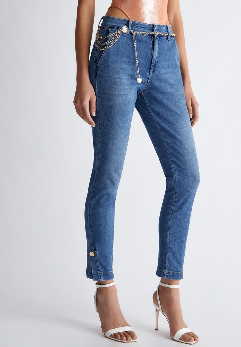 Femme portant un jean bleu slim avec une ceinture chaîne dorée et des sandales blanches à talons hauts à brides, debout devant un fond uni.