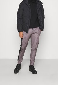 Graue gepolsterte Jacke, schwarzer Rollkragenpullover und gestreifte lila-graue Hose mit schwarzen Seitenpaneelen, kombiniert mit schwarzen Sneakers.